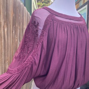 Puede incluir: Una blusa color ciruela con paneles de malla transparente y detalles florales bordados en las mangas. El escote redondo tambi&eacute;n est&aacute; adornado con bordados. Las mangas son largas y fruncidas en la mu&ntilde;eca, creando una forma voluminosa.