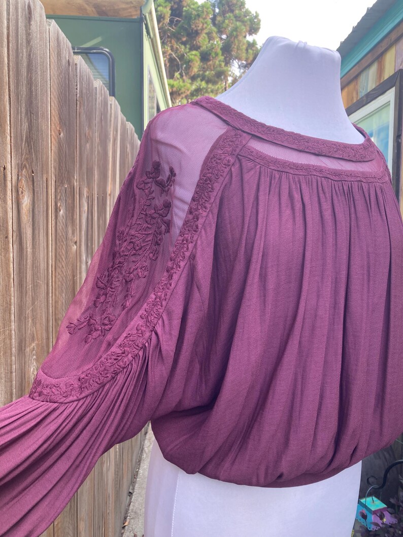 Puede incluir: Una blusa color ciruela con paneles de malla transparente y detalles florales bordados en las mangas. El escote redondo tambi&eacute;n est&aacute; adornado con bordados. Las mangas son largas y fruncidas en la mu&ntilde;eca, creando una forma voluminosa.
