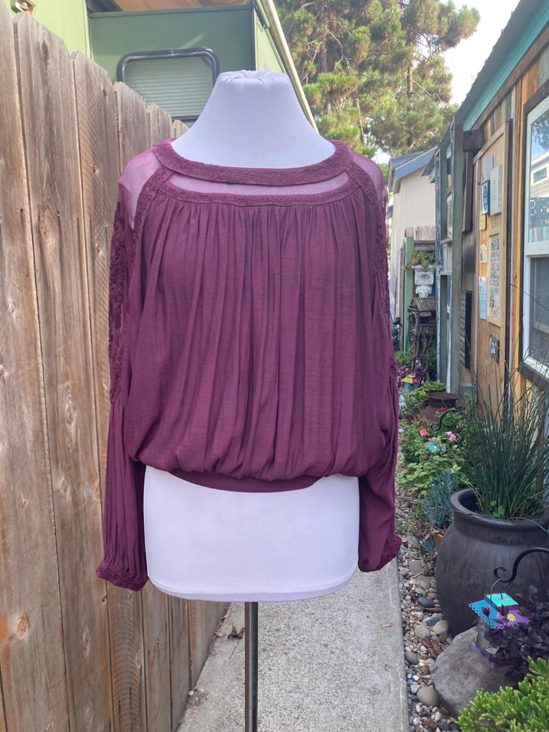 Puede incluir: Una blusa de manga larga color ciruela con cuello redondo y detalles de malla transparente. La blusa tiene cintura fruncida y detalles de encaje en las mangas. La parte superior se muestra en un maniqu&iacute; blanco.