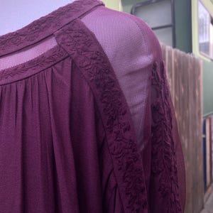Puede incluir: Una blusa color ciruela con paneles de hombros y mangas de malla transparente. El escote y las costuras de los hombros presentan detalles florales bordados. La tela tiene un aspecto texturizado, y la blusa se muestra en un maniqu&iacute;.