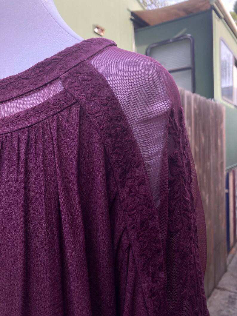 Puede incluir: Una blusa color ciruela con paneles de hombros y mangas de malla transparente. El escote y las costuras de los hombros presentan detalles florales bordados. La tela tiene un aspecto texturizado, y la blusa se muestra en un maniqu&iacute;.