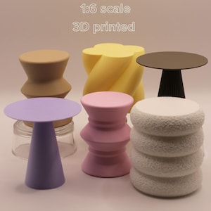 Könnte beinhalten: Eine Sammlung von 3D-gedruckten Miniaturtischen in verschiedenen Farben und Designs. Die Tische umfassen eine braune Sanduhrform, eine gelbe verdrehte Form, einen schwarz-braunen Tisch, einen lila Tisch, eine rosa Sanduhrform und einen weißen strukturierten Tisch. Der Text auf dem Bild lautet "1:6 scale 3D printed."