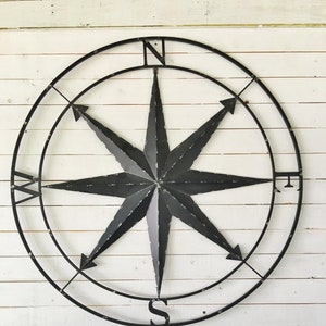 May include: Black metal compass with a star-shaped design and the cardinal directions N, S, E, and W. The compass is mounted on a white wooden wall.