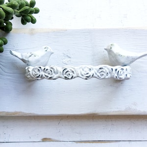 May include: White metal bird hook with a floral design. The hook is mounted on a white wooden wall.