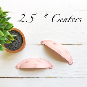 May include: Two pink metal cabinet pulls with a distressed finish. The pulls are 2.5 inches apart.