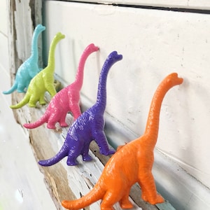 May include: Five colorful dinosaur figurines in a row on a weathered wooden surface. The dinosaurs are painted in shades of blue, green, pink, purple, and orange.