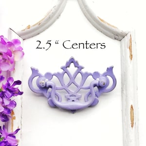May include: A lavender-colored metal cabinet pull with a decorative design. The pull has a 2.5 inch center to center measurement.