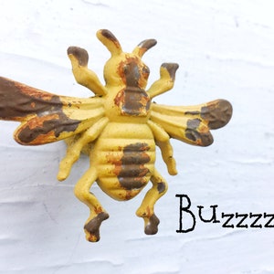 May include: A yellow and brown metal bee figurine with a distressed finish. The bee has wings, legs, and a stinger. The bee is mounted on a white wall. The text "Buzzzz..." is written below the bee.