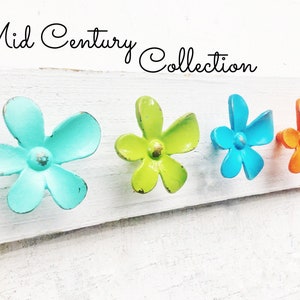 May include: Four metal flower-shaped hooks in turquoise, green, blue, and orange. The hooks are arranged in a row on a white wooden surface. The text "Mid Century Collection" is written above the hooks.