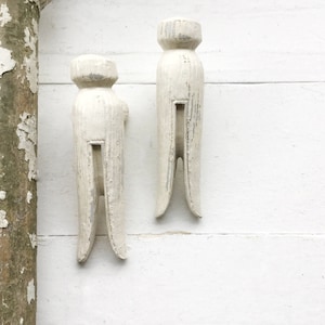 May include: Two white wooden clothespins, painted in a distressed white finish, are displayed against a white wood background. The clothespins are a vintage style and have a rustic look.