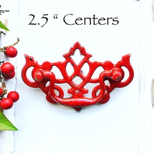 May include: Red metal drawer pull with an ornate design. The pull measures 2.5 inches between the centers.
