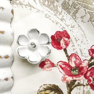 May include: White ceramic flower knob with a distressed finish. The knob is attached to a white fabric with a floral pattern.