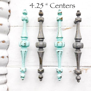 May include: Four antique-style cabinet knobs with a 4.25 inch center-to-center spacing. Two knobs are painted in a light turquoise blue with gold accents, and two knobs are painted in a dark gray with gold accents.