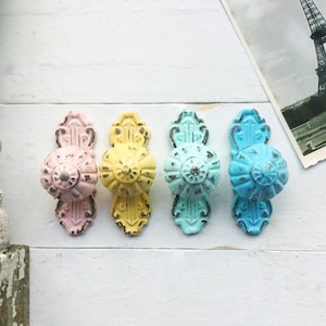 May include: Four decorative cabinet knobs in pastel colors. The knobs are in shades of pink, yellow, blue and green. Each knob has a detailed design and a distressed finish.
