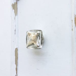 May include: A square glass cabinet knob with a silver finish. The knob is mounted on a white painted door with visible signs of wear and tear.