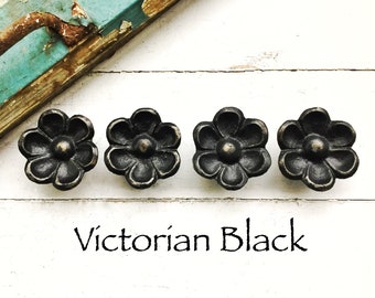May include: Four black Victorian-style flower-shaped cabinet knobs with a slightly raised center. The knobs are arranged in a row on a white wooden surface. The text "Victorian Black" is written below the knobs.