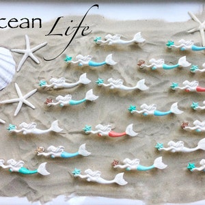 May include: A collection of white mermaid figurines with blue, turquoise, and coral tails. Each mermaid holds a star and is arranged on a sandy surface with white starfish and a seashell. The text "Ocean Life" is written at the top of the image.