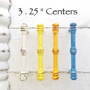 May include: Four colorful cabinet pulls in white, yellow, orange, and blue. Each pull is a vertical column with decorative rings and a distressed finish. The text "3.25" Centers" is above the pulls.