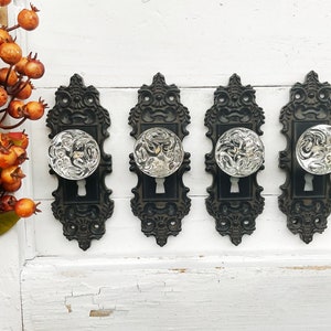 May include: Four black ornate door knobs with clear glass knobs. The knobs are mounted on a white wall.