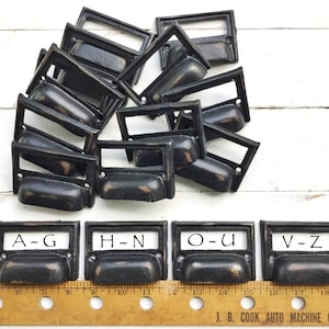 May include: Ten black metal drawer pulls with a label holder. The pulls are arranged in a fan shape. Four of the pulls have labels with the letters A-G, H-N, O-U, and V-Z. A ruler is in the bottom of the image.