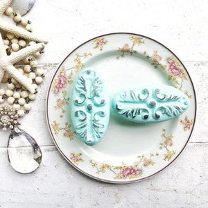 May include: Two oval-shaped decorative drawer pulls with a floral design. The pulls are painted a light blue color with silver accents. The pulls are displayed on a white plate with a floral pattern.