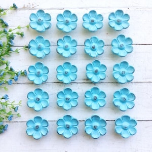 May include: A set of 18 blue flower-shaped cabinet knobs. The knobs are arranged in a grid pattern on a white wooden surface.