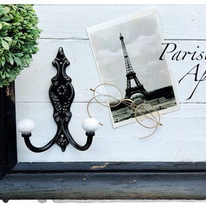 May include: A black metal double hook with white porcelain knobs mounted on a white wooden wall with a black frame. A vintage postcard of the Eiffel Tower and a pair of gold-rimmed glasses are on the wall. The text "Parisian Apt." is written in black cursive on the wall.
