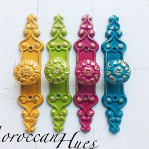 May include: Four ornate door knobs in yellow, green, pink, and blue with gold accents. The knobs are arranged in a row on a white background.