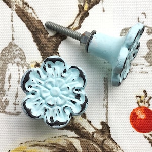 May include: Two light blue flower-shaped cabinet knobs with black accents. The knobs have a distressed finish and are made of metal.