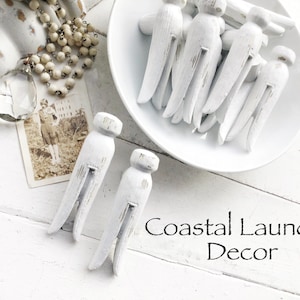 May include: White painted wooden clothespins with a distressed finish. The clothespins are arranged in a bowl and on a white wooden surface. The text "Coastal Laundry Decor" is written below the clothespins.