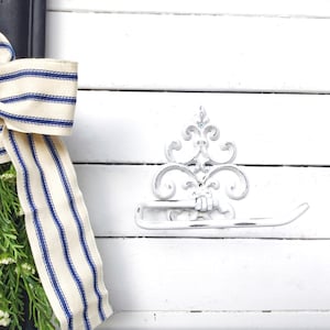 May include: White painted metal toilet paper holder with a decorative scroll design, mounted on a white wooden wall. A blue and white striped ribbon is tied around a bunch of white flowers.