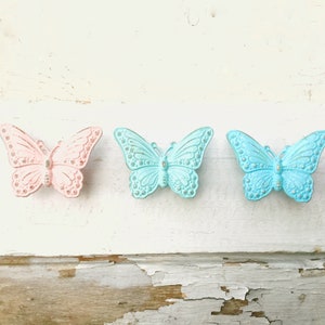 May include: Three decorative butterfly ornaments in pastel colors: pink, light blue, and blue. The butterfly wings have detailed patterns and small silver accents. The ornaments are displayed on a distressed white wooden surface.