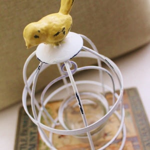 May include: White metal birdcage with a yellow bird perched on top. The birdcage is sitting on a book titled "Six Little Lambs of Aunt Eliza".