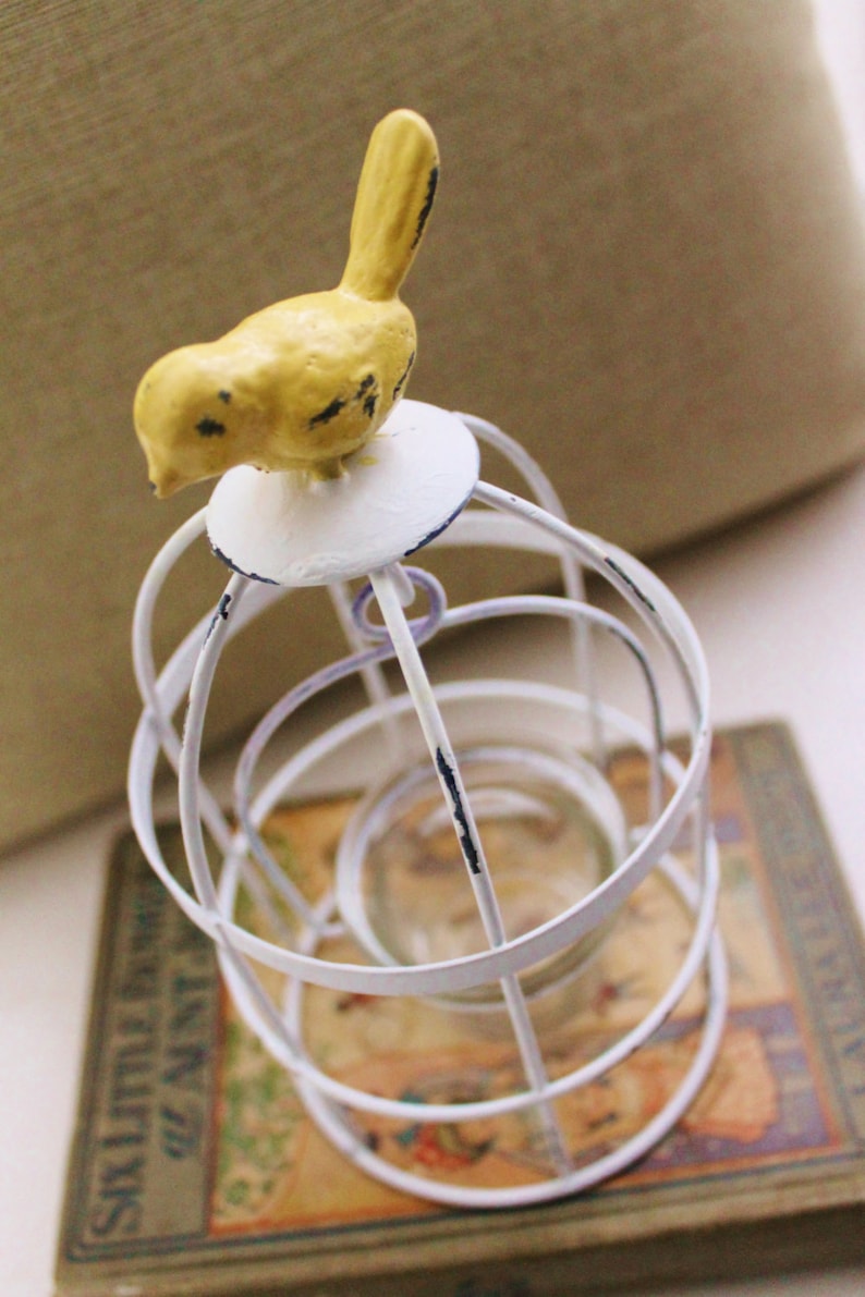 May include: White metal birdcage with a yellow bird perched on top. The birdcage is sitting on a book titled "Six Little Lambs of Aunt Eliza".