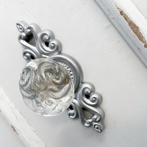 May include: A silver cabinet knob with a clear glass swirl design. The knob has a decorative, ornate base.