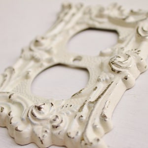 May include: Off-white, ornate light switch cover with a distressed finish. The cover features a decorative, baroque-style design with two circular openings. The edges and details have a subtle, metallic silver accent, adding depth to the design.