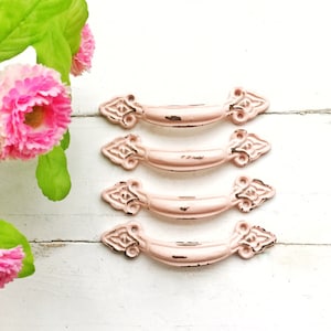 May include: Four pink metal drawer pulls with ornate detailing. The pulls are arranged in a row on a white wooden surface.