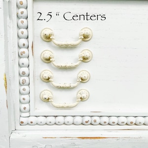 May include: Five white drawer pulls with a 2.5" center to center spacing. The pulls are arranged vertically on a white surface with a beaded trim.