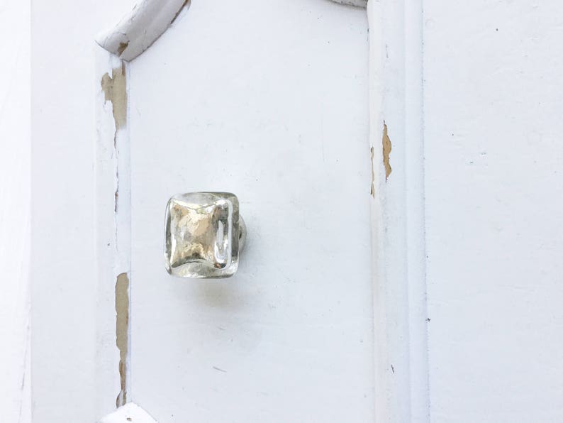 May include: A square glass cabinet knob with a silver finish. The knob is mounted on a white painted door with visible signs of wear and tear.