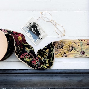 May include: A spool of beige ribbon, a black and white photograph, and gold-rimmed eyeglasses rest on a white surface. A decorative ribbon with floral patterns in red, yellow, and green is also present. The scene is framed by a black border.