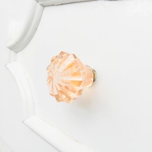 May include: A pale pink glass drawer knob with a faceted design. The knob is mounted on a white drawer front.