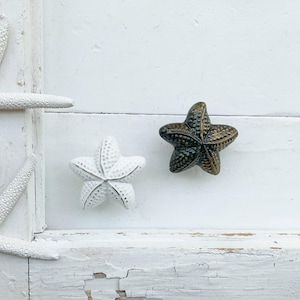 May include: Two ceramic starfish, one white and one brown, are mounted on a white painted wooden surface. The starfish are decorative and have a rustic look.