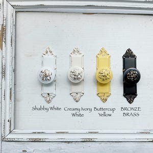 May include: Four decorative door knobs in a row, each with a different color and style. The colors are shabby white, creamy ivory white, buttercup yellow, and bronze brass. Each knob has a floral design and is mounted on a rectangular backplate. The text below each knob indicates the color.