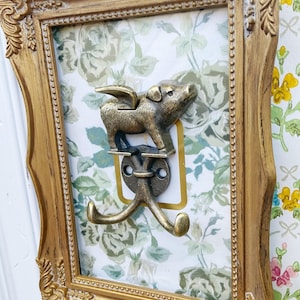 May include: A decorative wall hook featuring a bronze-colored flying pig design. The hook is mounted on a floral patterned background within a gold-colored ornate frame. The hook has two prongs for hanging items.