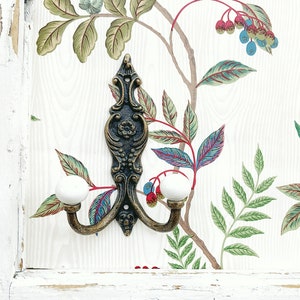 May include: A wall-mounted coat hook featuring an ornate, dark bronze-toned metal backplate with intricate scrollwork and a central floral motif. Two curved arms extend from the base, each tipped with a smooth, round, white ceramic knob.