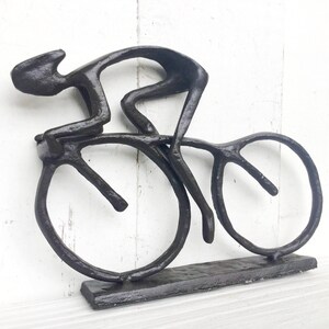 May include: A black metal sculpture of a cyclist riding a bicycle. The sculpture is abstract and stylized, with the cyclist's body and the bicycle's wheels represented by simple lines.
