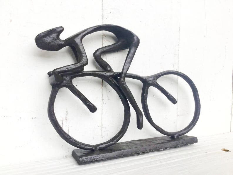 May include: A black metal sculpture of a cyclist riding a bicycle. The sculpture is abstract and stylized, with the cyclist's body and the bicycle's wheels represented by simple lines.