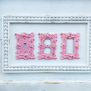 May include: Three decorative light switch covers in a white frame with a beaded border. The covers are pink with gold accents and ornate designs.