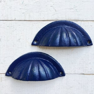 May include: Two blue cast iron drawer pulls with a scalloped design. The pulls are on a white wooden surface.