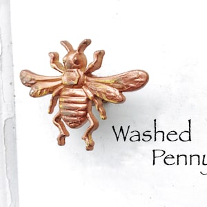 May include: A gold bee-shaped cabinet knob with a washed penny finish. The knob is on a white background with the text "Washed Penny" below it.